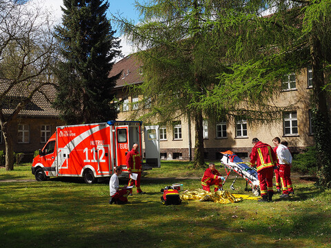 Rettungswagen mit Einsatzkräften auf Wiese unter Bäumen bei sonnigem Wetter